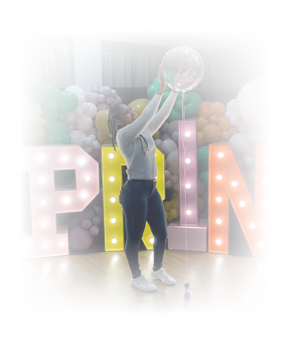 Jeune femme souriante levant un ballon transparent sous des lettres lumineuses formant 'PAIN' et un mur de ballons pastel lors d'une fête.