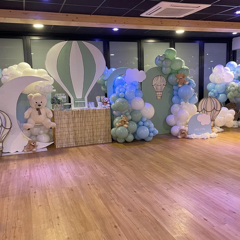 Décoration de fête sur le thème montgolfière et oursons en peluche, avec ballons bleus, verts et blancs, et un panneau "MELVYN JAN".