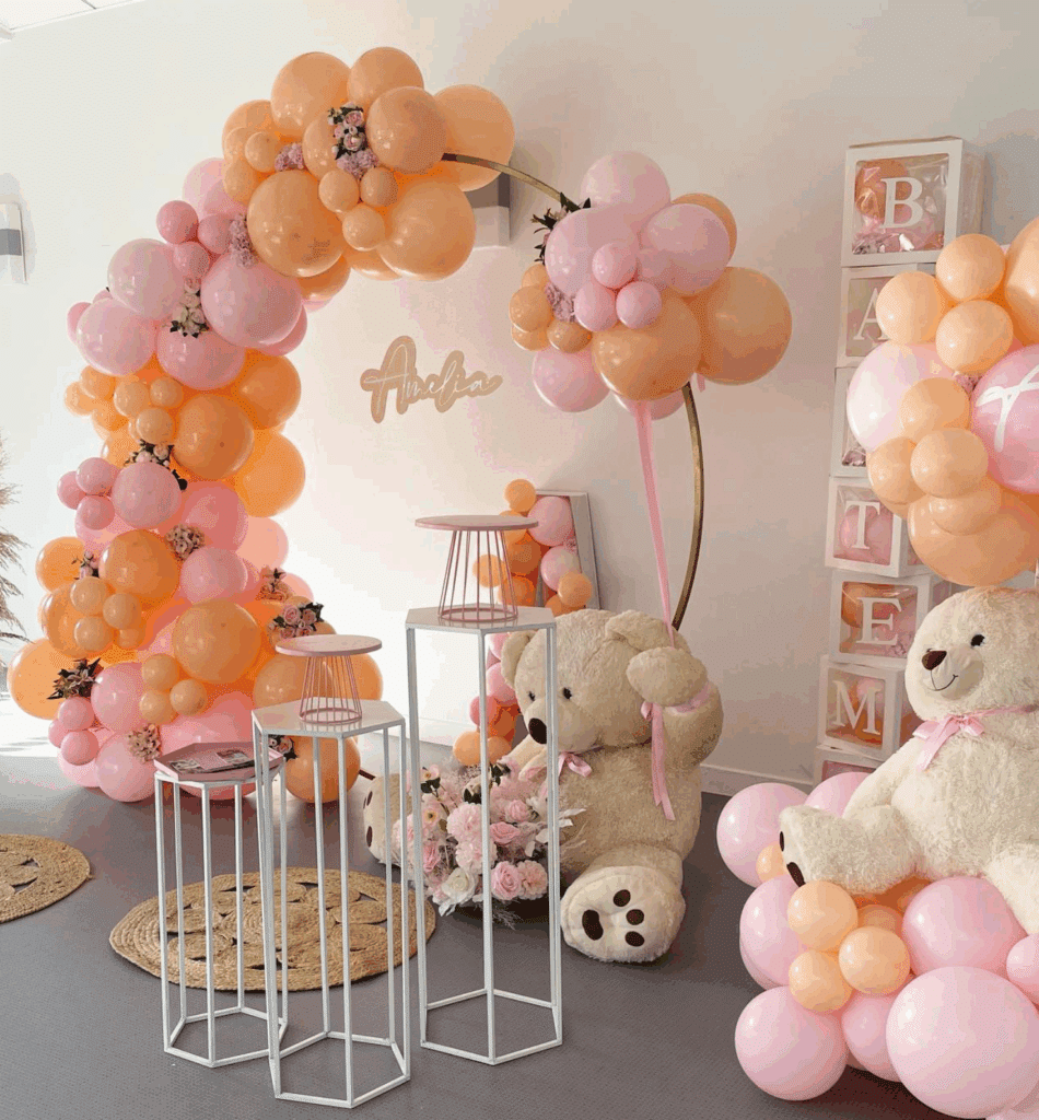 Décoration de fête pastel avec arche de ballons rose et pêche, inscription 'Amelia', grands ours en peluche et présentoirs blancs.